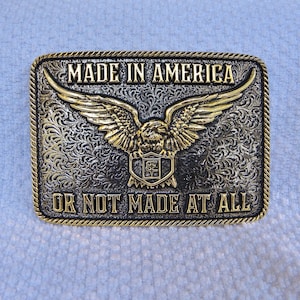 May include: Rectangular metal belt buckle with a gold eagle design and the text "MADE IN AMERICA OR NOT MADE AT ALL". The buckle has a decorative border and a detailed floral pattern background.