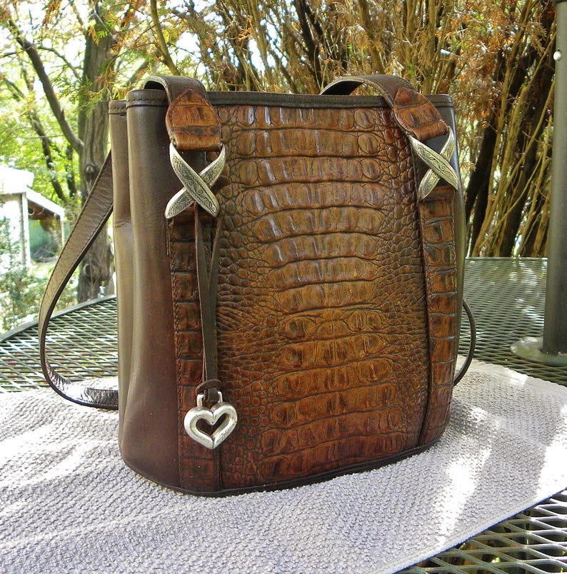 May include: A brown leather tote bag with a crocodile-textured front panel. The bag features a shoulder strap, decorative silver accents, and a heart-shaped charm. The bag is sitting on a white towel.