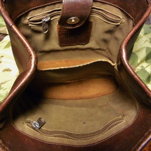 May include: Open view of a brown leather handbag with a top flap and a magnetic closure. The interior features multiple compartments, including zippered pockets and a main storage area. The bag has a classic, vintage aesthetic.