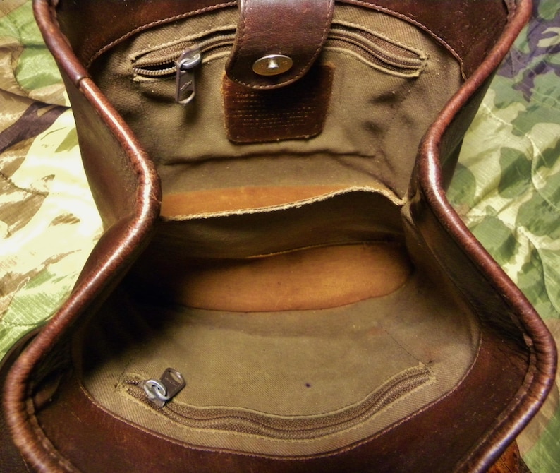 May include: Open view of a brown leather handbag with a top flap and a magnetic closure. The interior features multiple compartments, including zippered pockets and a main storage area. The bag has a classic, vintage aesthetic.