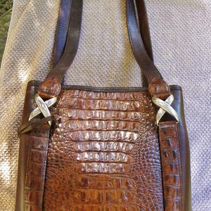 May include: Brown leather tote bag with a crocodile skin pattern. The bag has two dark brown leather straps and silver-colored X-shaped hardware. The bag is sitting on a light brown towel.