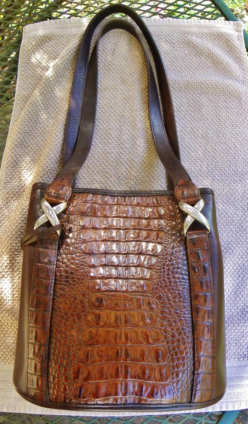May include: Brown leather tote bag with a crocodile skin pattern. The bag has two dark brown leather straps and silver-colored X-shaped hardware. The bag is sitting on a light brown towel.