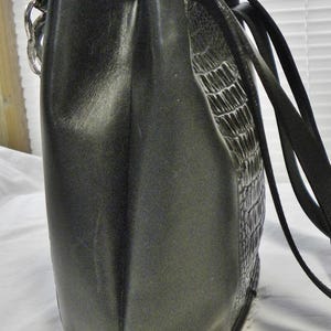 May include: A black handbag with a textured, crocodile-like pattern on one side. The bag has a smooth, matte finish on the other side and a black strap. The bag is rectangular with a rounded bottom.