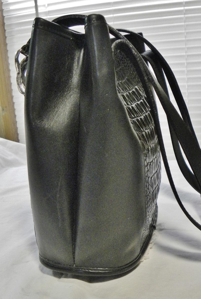 May include: A black handbag with a textured, crocodile-like pattern on one side. The bag has a smooth, matte finish on the other side and a black strap. The bag is rectangular with a rounded bottom.