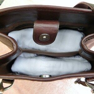 May include: A brown leather handbag with a top handle and a magnetic snap closure. The interior is visible, revealing a zippered pocket and two white, padded inserts. The bag has silver-tone hardware.