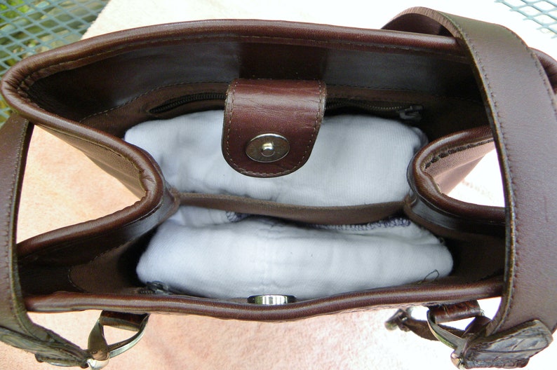 May include: A brown leather handbag with a top handle and a magnetic snap closure. The interior is visible, revealing a zippered pocket and two white, padded inserts. The bag has silver-tone hardware.