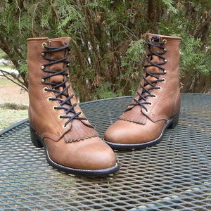 May include: A pair of brown leather boots with black laces and a fringe detail on the toe. The boots have a mid-calf height and are set on a black sole with a white stitch.