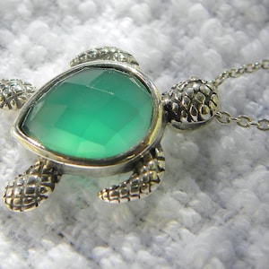 May include: A silver-toned turtle pendant necklace. The turtle features a large, faceted, emerald-green gemstone set in a silver frame. The turtle's shell, head, and feet have a textured, scale-like design. The necklace has a delicate chain.