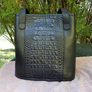 May include: Black tote bag with a faux crocodile skin center panel and smooth black sides. The bag has two short handles and a rectangular shape. The bag is sitting on a pinkish-tan surface.