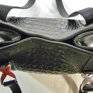 May include: Black leather handbag with a textured, crocodile-like pattern. The bag has a unique, open design with two handles and decorative metal accents. The bag's straps are black.