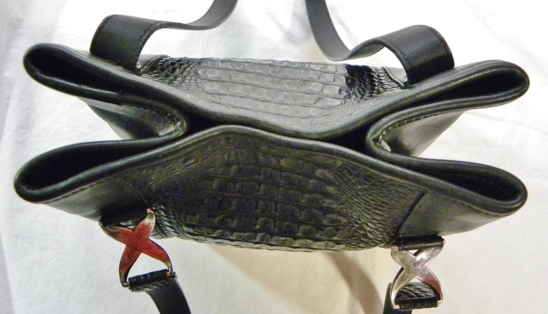 May include: Black leather handbag with a textured, crocodile-like pattern. The bag has a unique, open design with two handles and decorative metal accents. The bag's straps are black.