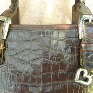 May include: Close-up of a brown leather handbag with a crocodile skin texture. The bag features silver buckles and a heart-shaped charm. The braided handle and the bag's overall design suggest a vintage or luxury aesthetic.