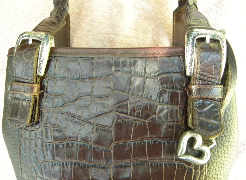 May include: Close-up of a brown leather handbag with a crocodile skin texture. The bag features silver buckles and a heart-shaped charm. The braided handle and the bag's overall design suggest a vintage or luxury aesthetic.