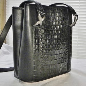 May include: Black shoulder bag with a faux crocodile skin texture on the front panel. The bag has a long strap, silver-toned hardware, and a structured design. The bag is made of a smooth, matte material.