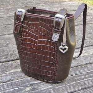 May include: A brown leather handbag with a crocodile-textured front panel. The bag features silver-tone buckles, a braided strap, and a heart-shaped charm. The bag is a bucket style with a structured shape.