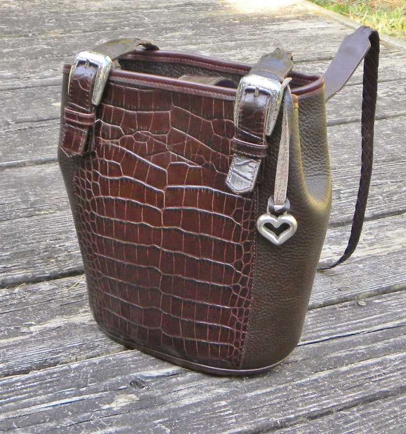 May include: A brown leather handbag with a crocodile-textured front panel. The bag features silver-tone buckles, a braided strap, and a heart-shaped charm. The bag is a bucket style with a structured shape.