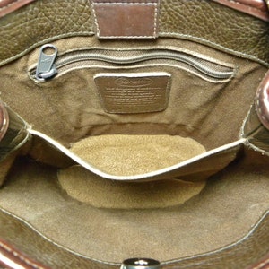 May include: Open view of a brown leather handbag interior. The bag features a zippered pocket, a small interior pocket, and a metal tag. The leather has a textured appearance, and the interior lining is a lighter brown color.