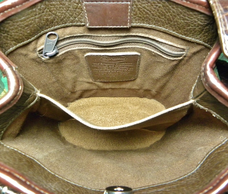 May include: Open view of a brown leather handbag interior. The bag features a zippered pocket, a small interior pocket, and a metal tag. The leather has a textured appearance, and the interior lining is a lighter brown color.
