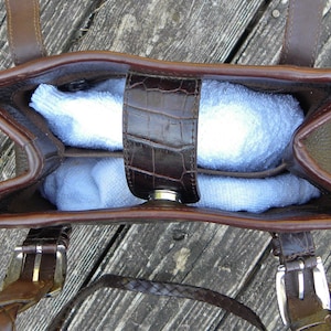 May include: A brown leather handbag with a top handle and a shoulder strap. The bag has a crocodile-embossed flap and a silver clasp. The interior of the bag is filled with a light blue item. The bag is sitting on a wooden surface.