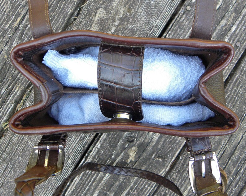 May include: A brown leather handbag with a top handle and a shoulder strap. The bag has a crocodile-embossed flap and a silver clasp. The interior of the bag is filled with a light blue item. The bag is sitting on a wooden surface.