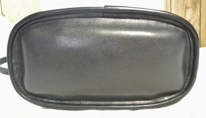 May include: A black leather handbag with a rounded rectangular base. The bag has a smooth, matte finish and a contrasting black trim around the edges. The image is a close-up of the bottom of the bag.