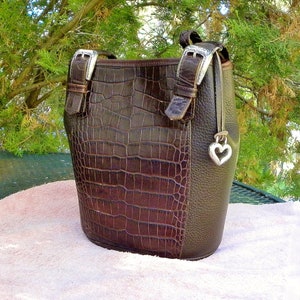 May include: A brown handbag with a crocodile-textured front panel and a smooth, dark brown side panel. The bag features silver-colored buckles and a heart-shaped charm. The bag has a handle and a shoulder strap.