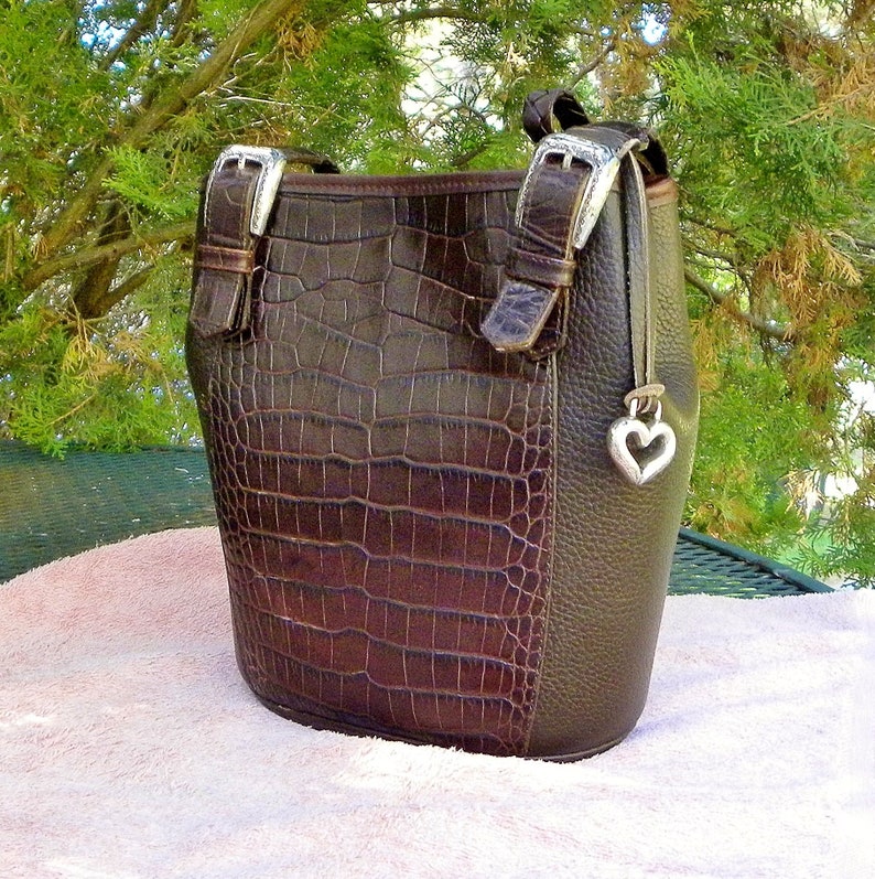 May include: A brown handbag with a crocodile-textured front panel and a smooth, dark brown side panel. The bag features silver-colored buckles and a heart-shaped charm. The bag has a handle and a shoulder strap.