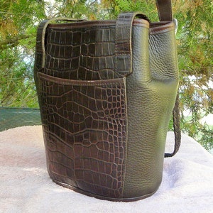 May include: A dark brown leather handbag with a crocodile-textured front panel and a smooth, textured side panel. The bag has a shoulder strap and a braided handle. The bag is sitting on a white surface.