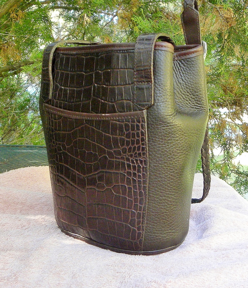 May include: A dark brown leather handbag with a crocodile-textured front panel and a smooth, textured side panel. The bag has a shoulder strap and a braided handle. The bag is sitting on a white surface.