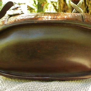 May include: A brown leather handbag with a rounded bottom and a textured top. The bag has a shoulder strap and silver-colored hardware. The leather appears to have a glossy finish, and the bag is sitting on a textured surface.