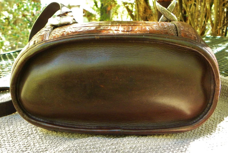 May include: A brown leather handbag with a rounded bottom and a textured top. The bag has a shoulder strap and silver-colored hardware. The leather appears to have a glossy finish, and the bag is sitting on a textured surface.