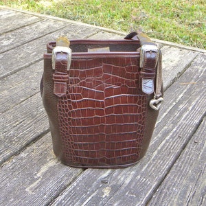 May include: A brown leather handbag with a crocodile-textured front panel. The bag has a bucket shape, silver buckles, and a small heart charm. The handle is a dark brown color. The bag is sitting on a wooden surface.