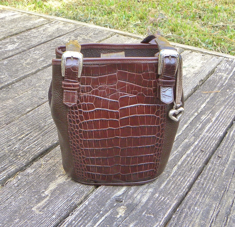 May include: A brown leather handbag with a crocodile-textured front panel. The bag has a bucket shape, silver buckles, and a small heart charm. The handle is a dark brown color. The bag is sitting on a wooden surface.
