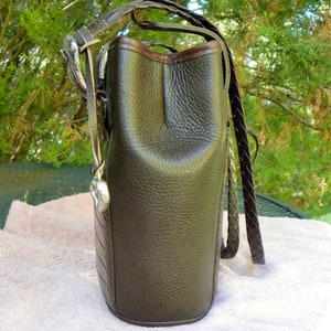 May include: A dark brown leather handbag with a textured finish. The bag features a braided strap and a silver-toned metal accent. The top edge is trimmed with a darker brown leather. The bag has a heart-shaped charm.