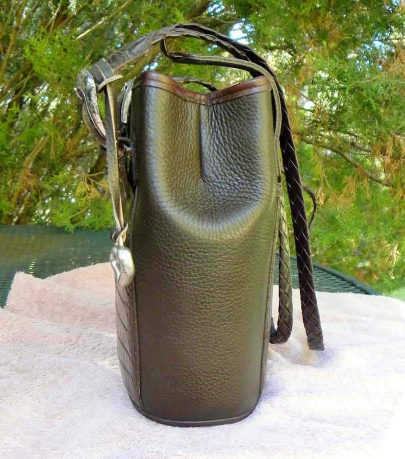 May include: A dark brown leather handbag with a textured finish. The bag features a braided strap and a silver-toned metal accent. The top edge is trimmed with a darker brown leather. The bag has a heart-shaped charm.
