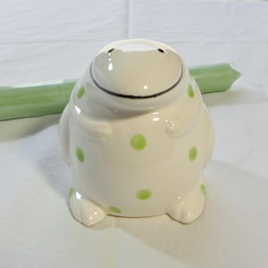 May include: A ceramic figurine of a smiling, round creature with a white base and green polka dots. The figurine has small black eyes and a black curved line for a mouth. A green candle is in the background.