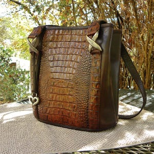 May include: Brown leather handbag with a crocodile skin pattern. The bag has a shoulder strap and decorative metal accents. A heart-shaped charm hangs from the side. The bag is sitting on a tan surface.
