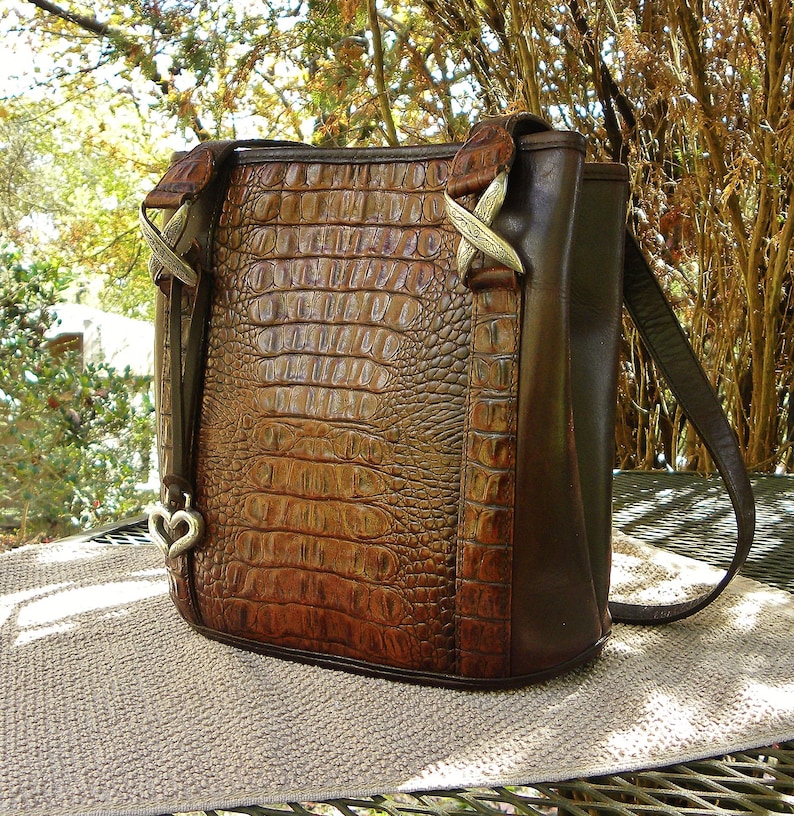 May include: Brown leather handbag with a crocodile skin pattern. The bag has a shoulder strap and decorative metal accents. A heart-shaped charm hangs from the side. The bag is sitting on a tan surface.