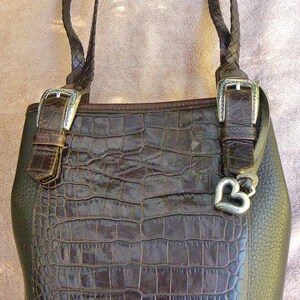 May include: A brown leather handbag with a crocodile-skin texture on the front. The bag has braided handles, silver buckles, and a silver heart charm. The bag is approximately 12 inches tall.