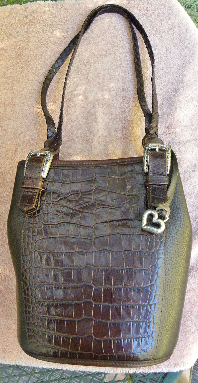 May include: A brown leather handbag with a crocodile-skin texture on the front. The bag has braided handles, silver buckles, and a silver heart charm. The bag is approximately 12 inches tall.