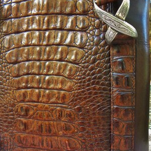 May include: Close-up of a brown leather bag with a crocodile skin texture. The bag features a silver-colored decorative clasp. The leather has a rich, textured appearance, with varying shades of brown. The bag's design suggests a focus on craftsmanship and luxury.