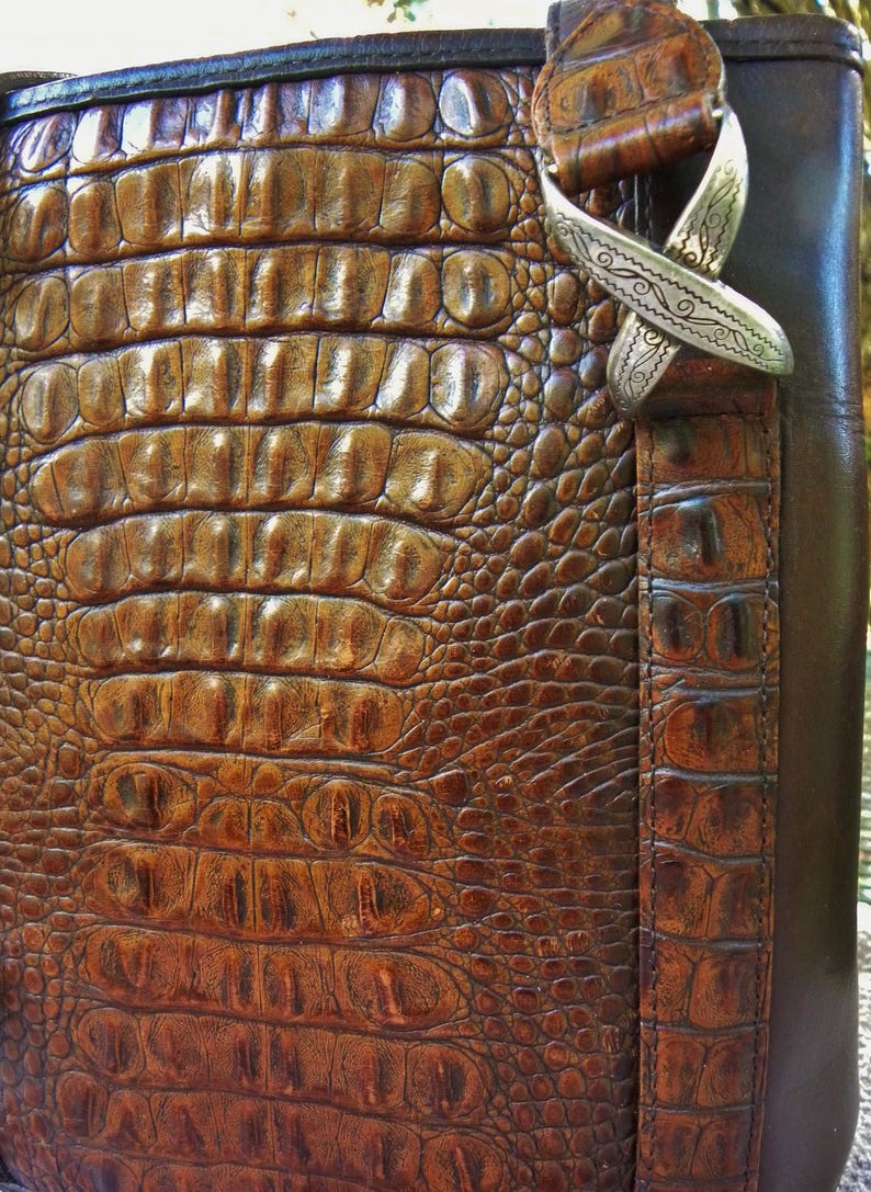 May include: Close-up of a brown leather bag with a crocodile skin texture. The bag features a silver-colored decorative clasp. The leather has a rich, textured appearance, with varying shades of brown. The bag's design suggests a focus on craftsmanship and luxury.