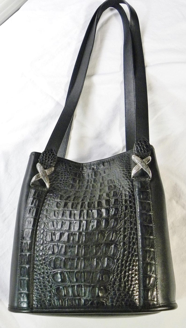 May include: Black leather tote bag with a crocodile-textured center panel. The bag features two shoulder straps and silver-colored decorative accents. The bag is designed with a structured shape and a classic, elegant style.