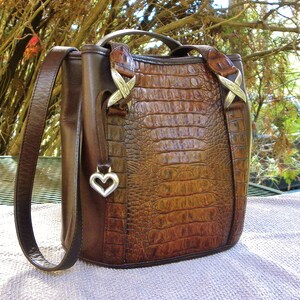May include: A brown leather handbag with a crocodile-textured front panel. The bag has a top handle, a shoulder strap, and silver-toned hardware. A silver heart-shaped charm hangs from the side. The bag is approximately 10 inches tall.