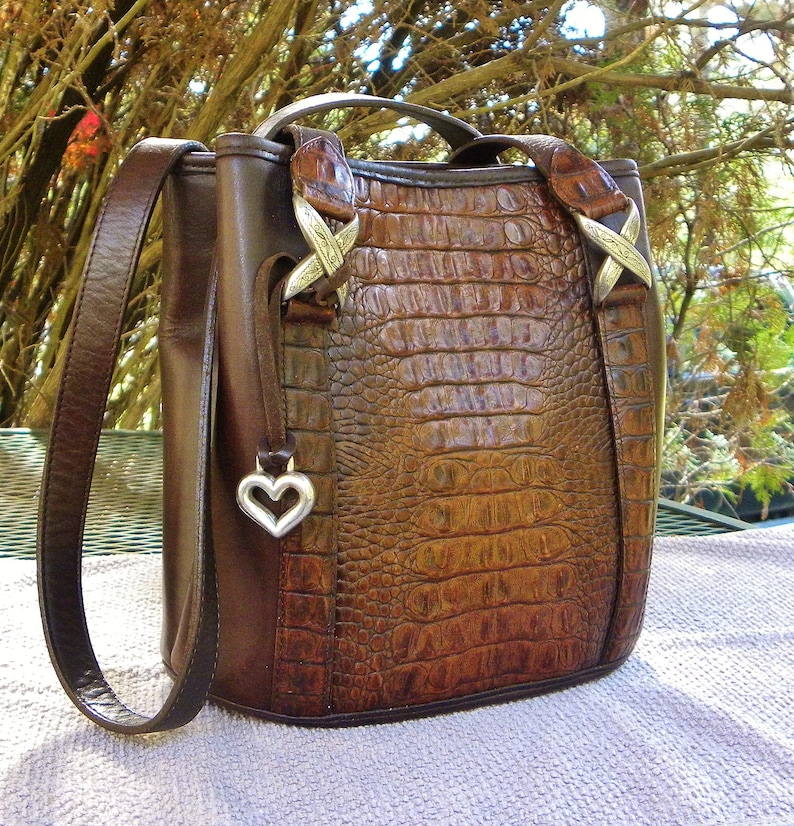 May include: A brown leather handbag with a crocodile-textured front panel. The bag has a top handle, a shoulder strap, and silver-toned hardware. A silver heart-shaped charm hangs from the side. The bag is approximately 10 inches tall.