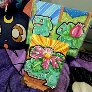 Peut inclure: Trois illustrations colorées de Bulbasaur et de ses évolutions sont présentées. L'œuvre présente des teintes vertes, roses et jaunes vibrantes, sur des fonds de ciels bleus et de champs herbeux. Les dessins sont dans des pochettes de protection.
