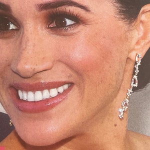 May include: A close-up of a woman's face with a smile. She is wearing a pair of long, dangling earrings with small, clear stones.