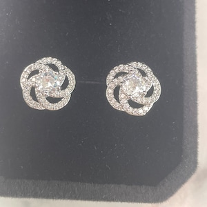 May include: A pair of silver-coloured stud earrings. Each earring has a central, clear, round stone surrounded by smaller, clear stones in a floral design. The earrings are displayed on a black velvet surface.