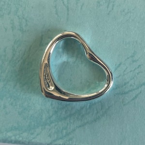 Puede incluir: Un colgante de corazón de plata. El colgante está abierto y tiene un acabado liso y pulido.