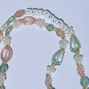 May include: A beaded accessory with the text "MRS YOUNG" in gold lettering. The beads are in shades of pink, green, and clear, with flower and teardrop shapes. The beads are strung on a gold-colored chain.
